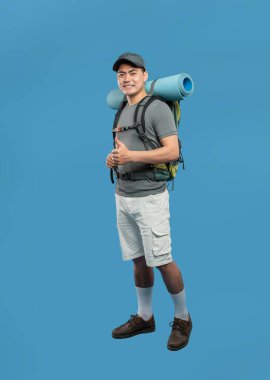 Sırt çantası ve mavi bir uyku paspası olan gülümseyen bir adam mavi bir arka planda durur.