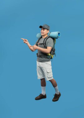 Sırt çantalı neşeli bir yürüyüşçü mavi arka planda duruyor. Görülmemiş bir yere bakıyor.