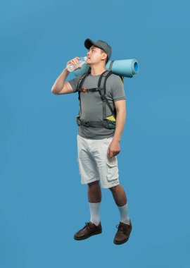 Asyalı bir erkek yürüyüşçü, mavi arka planda yürüyüş kıyafeti giyerek su içerken görülüyor.