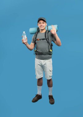 Gülümseyen Asyalı bir erkek yürüyüşçü bir şişe sırt çantasıyla maceraya hazır ve maviliğe karşı yuvarlanmış bir paspas.