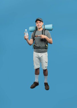 Asyalı bir erkek yürüyüşçü sırt çantası şort şapkası takıp gülümsüyor ve elinde bir su şişesi tutuyor.