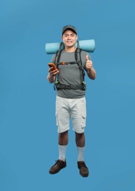 Sırt çantası ve uyku paspası olan bir adam cep telefonunu tutarken başparmağını kaldırıyor. Mutlu yürüyüşçü.