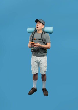 Yürüyüş kıyafeti giymiş bir adam cep telefonuyla mavi bir arka plana karşı sırt çantası takıyor.