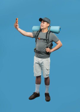 Sırt çantalı bir yürüyüşçünün mavi arka planda günlük kıyafetlerle selfie çekerken tam bir fotoğrafı.