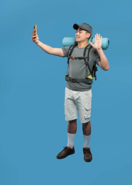 Yürüyüş kıyafeti giymiş bir adam telefonunu açık hava macerasına hazır mavi arka planda kullanır.