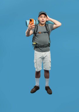 Sırt çantası ve uyku paspası olan sürpriz bir yürüyüşçü mavi bir arkaplanda selfie çeker.