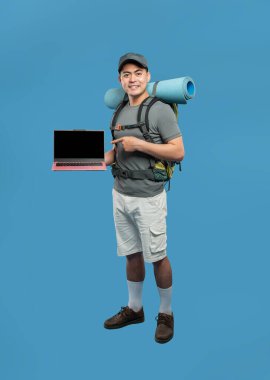 Sırt çantasıyla gülümseyen yürüyüşçü mavi bir arkaplanda sunar ve boş bir dizüstü bilgisayara işaret eder