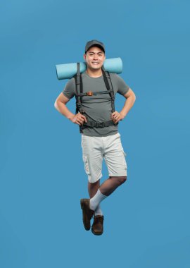 Gülümseyen bir adam mavi bir arka plana atlıyor. Sırt çantası şort şapkası ve günlük kahverengi ayakkabılar giyiyor.