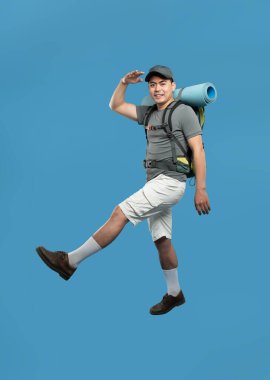 Sırt çantası şapkalı ve uyku tulumlu bir adam öne doğru yürür ve mavi arka plana bakar.