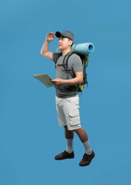 Sırt çantası ve haritası olan bir adamın tam görüntüsü. Saf mavi bir arkaplan ile uzaklığa bakıyor.