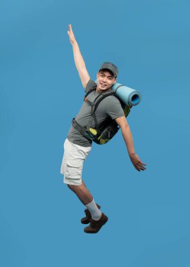 Yürüyüş kıyafetleri olan neşeli bir adam mavi arka planda, açık hava macerasının önünde mutlu bir şekilde sıçrıyor.