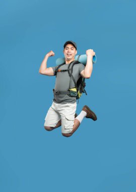 Enerjik bir erkek yürüyüşçü havada sıçrıyor sırt çantasıyla kaslarını esnetiyor ve mavi arka plana doğru yatıyor.