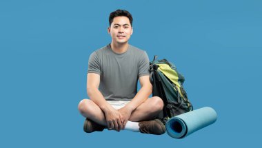 Sırt çantası ve uyku paspası ile bağdaş kurmuş genç adam yolculuk için hazır bekliyor.