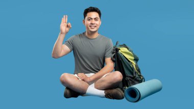 Neşeli bir yürüyüşçü iyi bir el hareketi yapar. Yanında bir sırt çantası ve yoga minderi vardır.