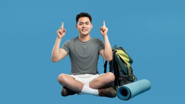 Genç adam mavi bir arka planın önünde bağdaş kurmuş oturuyor. Dışarıdaki kıyafetleri gösteriyor.