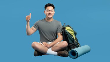 Gülümseyen gezgin sırt çantasıyla dinleniyor ve mavi arka planda baş parmak hareketi yapıyor.