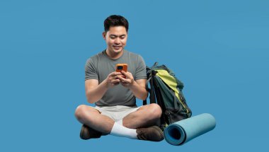 Sırt çantası ve yoga minderiyle oturan bir adam telefonuna gülümsüyor.