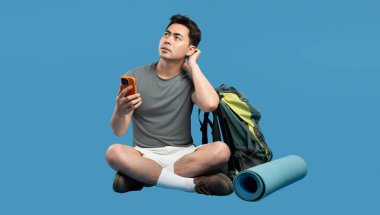 Bir adam elinde telefon, elinde yürüyüş teçhizatıyla oturur ve dikkatlice mavi üzerine bakar.