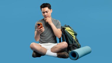 Kafası karışmış bir adam sırt çantası ve minderle mavi arka plandaki telefona bakıyor. Teknoloji ve seyahat.