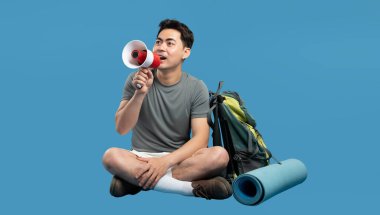 Sırt çantalı ve yoga minderli bir megafonla mavi arka planda oturan tutkulu bir gezgin.