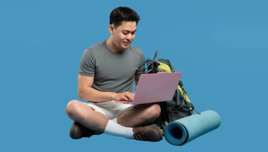 Asyalı bir adam dizüstü bilgisayarla sırt çantası ve yoga minderiyle bacak bacak bacak üstüne atarak oturur.