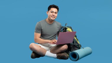 Asyalı bir adam dizüstü bilgisayarla seyahat planlıyor. Yanında bir sırt çantası ve yoga minderi var.