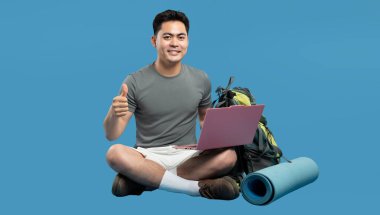 Gülümseyen gezgin dizüstü bilgisayarlı sırt çantası ve paspas karıştırma teknolojisiyle mavi üzerine açık hava macerası ile oturur.