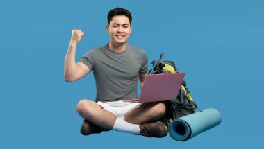 Adam sırt çantası ve uyku tulumuyla oturur ve seyahat planlarını yumruğunu kaldırarak kutlar.