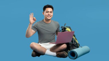 Asyalı bir adam sırt çantası ve paspasla dizüstü bilgisayarla bacak bacak bacak üstüne atarak oturuyor ve 