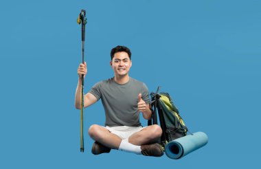 Yürüyüş teçhizatıyla gülümseyen bir yürüyüşçü, temiz bir atış için sağlam mavi bir arkaplana karşı başparmak kaldırır