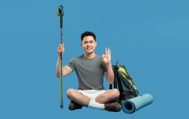 Gülümseyen yürüyüşçü teçhizat ile çapraz oturur: Sırt çantası yürüyüş direkleri ve sert mavi bir zemin üzerinde mat