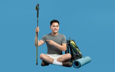 Asyalı bir adam yürüyüş takımlarıyla oturur. Sırık çantası ve paspas da dahil. Mavi bir stüdyo arka planında.