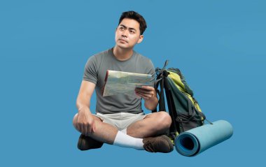 Yanında bir seyahat çantası planlayan adam mavi bir arka planda seyahat etmeden önce bir harita üzerinde çalışıyor.