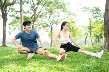 Genç bir çift açık havada parkta egzersiz yapıyor. Aydınlık bir günde bacaklarını açıyorlar.