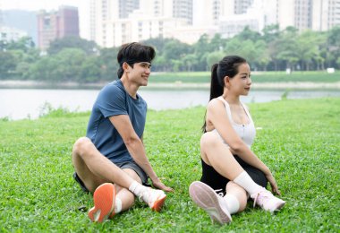 Atletik giysiler içinde şehir ve su arka planıyla çimlerin üzerinde uzanan bir çift.