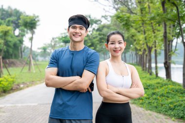 Gülümseyen bir erkek ve kadın spor kıyafetleri içinde kolları çaprazlanmış bir şekilde parkta poz veriyorlar.