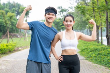 Spor kıyafetleri içinde gülümseyen bir çift, sağlık ve zindelik saçan bir parkta kaslarını esnetiyor.