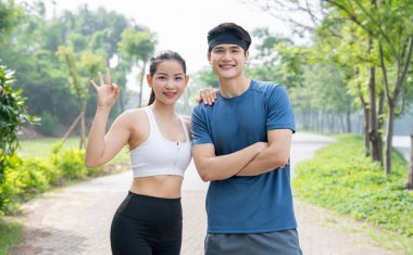 Atletik giyinmiş iki gülen insan, ağaçlar ve yeşilliklerle çevrili bir parkta poz veriyorlar. Mutlular.