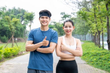 Mutlu genç adam ve kadın ağaçlık bir yolda ferahlatıcı bir egzersizden sonra başparmaklarını kaldırıyorlar.
