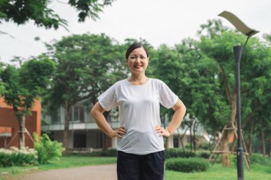 Atletik giyinmiş gülümseyen bir kadın yemyeşil bir parkta poz veriyor ve sağlık ve canlılık yayıyor.