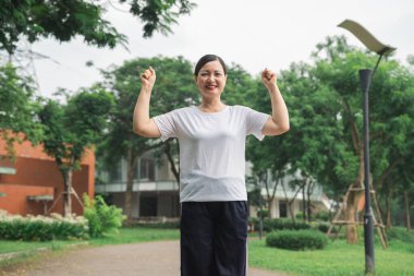 Beyaz gömlekli ve siyah pantolonlu gülümseyen bir kadın parkta kollarını kaldırıyor. Mutlu görünüyor.