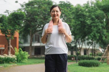 Gülümseyen bir kadın dışarıda egzersiz yaptıktan sonra boynunda havluyla parkta dikiliyor.