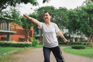 Bir kadın dışarıda, sakin bir parkta fiziksel ve ruhsal sağlığını geliştirmek için Tai Chi alıştırması yapıyor.