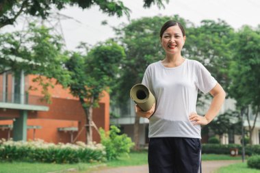 Elinde yoga minderi olan gülümseyen bir kadın yeşil ve sakin bir ortamda açık havada egzersiz yapmaktan hoşlanır.