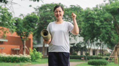 Dışarıda yoga minderiyle çalışmaya hazır bir kadın gülümsemesi ve parkta kaldırılmış bir kolla.