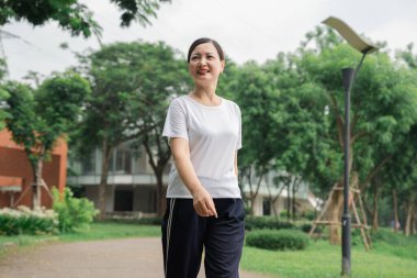 Parkta beyaz bluzlu ve siyah pantolonlu bir kadın gülümsüyor.