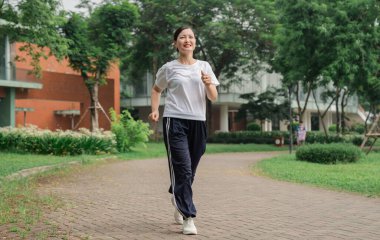 Tuğla yolda koşan antrenman kıyafetli gülümseyen bir kadın. Etrafı ağaçlarla çevrili ve parkta yeşillik var.