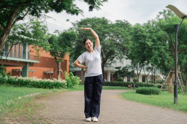 Beyaz gömlekli ve siyah pantolonlu bir kadın parkta esniyor. Güneşli bir gün.