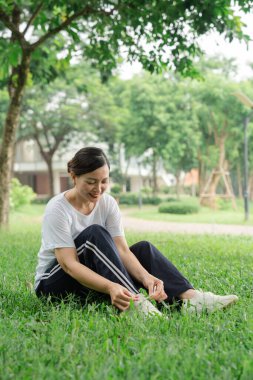 Bir kadın çimenlerde oturmuş ayakkabı bağcıklarını bağlıyor. Manzaralı parkta egzersiz yapmaya hazırlanıyor.