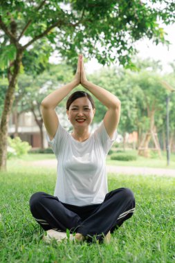 Spor kıyafetleri içinde gülümseyen bir kadın açık havada yoga yapıyor yemyeşil bir parkta sağlık için meditasyon yapıyor.